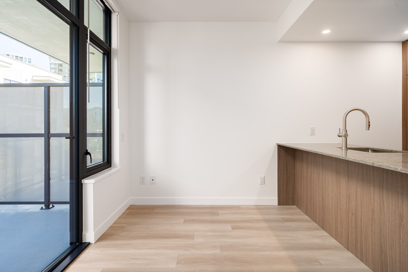 A modern, empty apartment interior featuring light wood flooring, a kitchen area with a marble countertop, and sliding glass doors leading to a balcony.