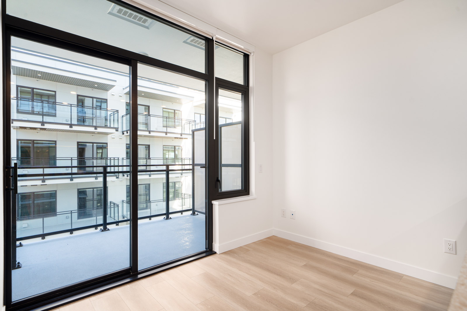 Unfurnished room with light wood flooring, white walls, and large glass doors leading to a balcony overlooking a modern apartment complex.