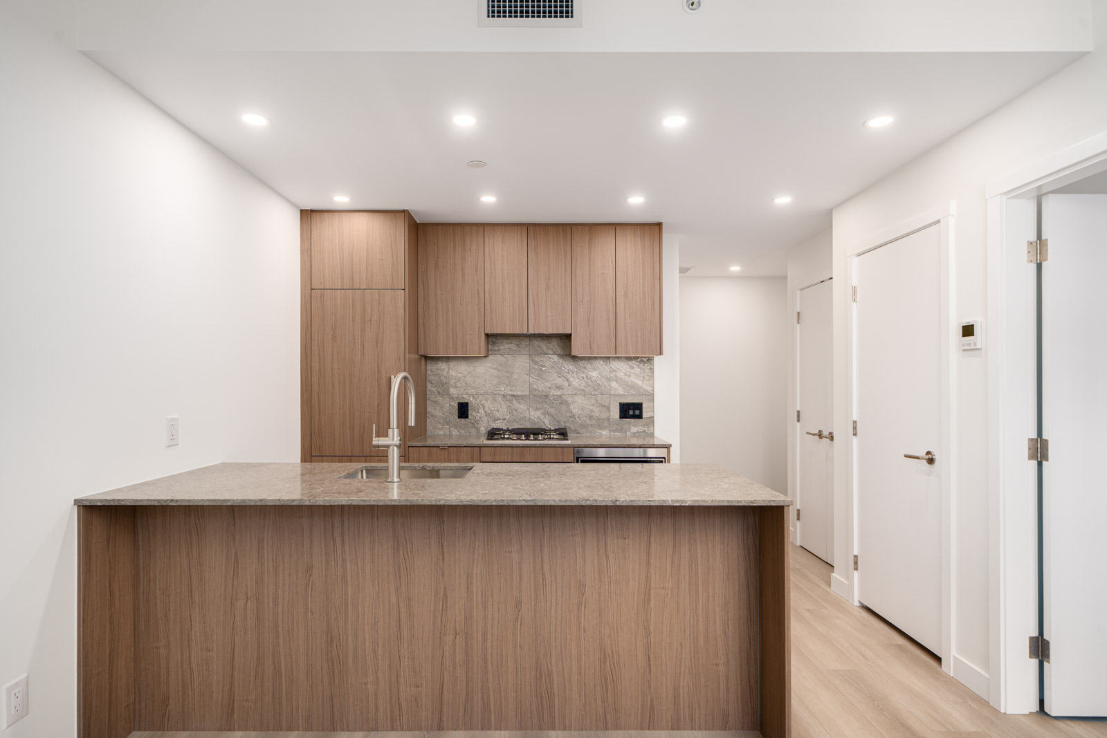 Modern kitchen with wood cabinets, a stone countertop and backsplash, built-in appliances, and recessed ceiling lights.