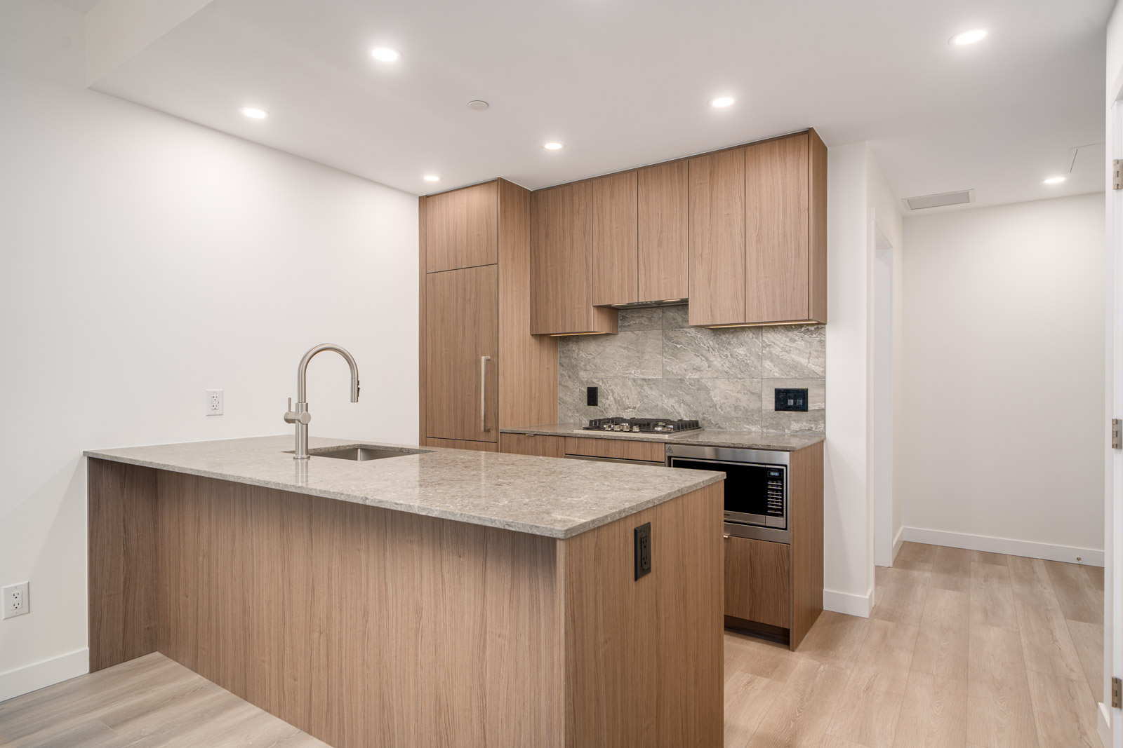 Modern kitchen with wood cabinetry, marble countertops, built-in stove, sink on island, integrated appliances, and recessed ceiling lighting.