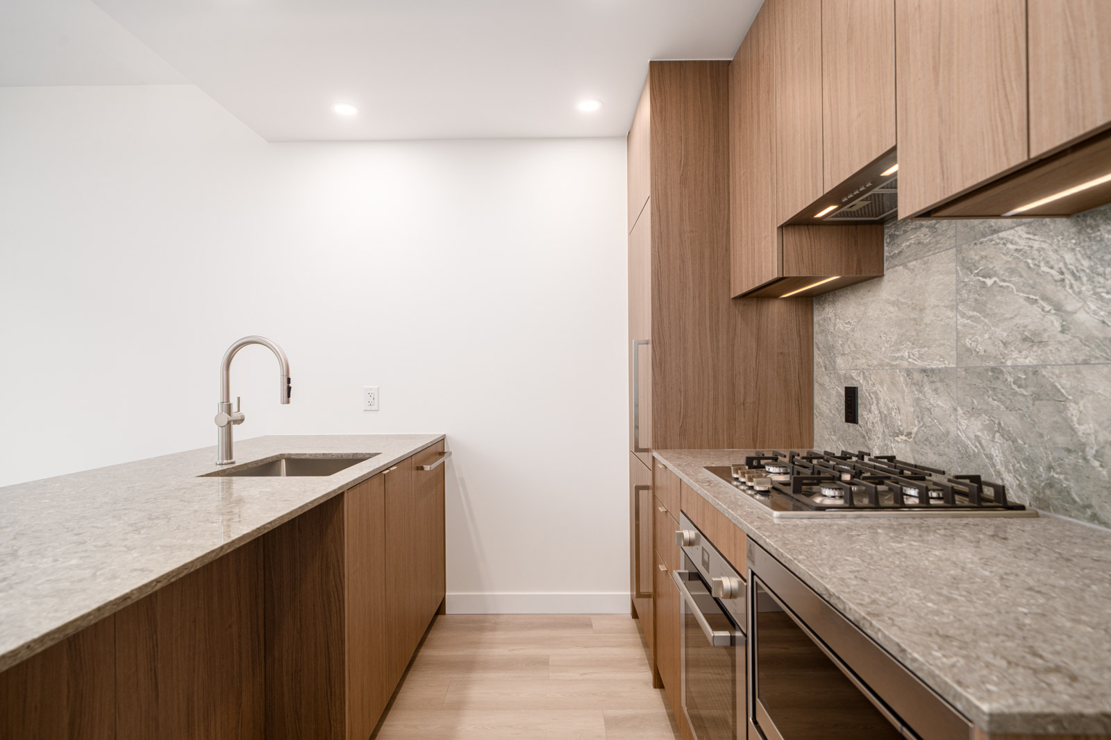 Modern kitchen with wood cabinets, marble countertops, built-in oven, gas cooktop, integrated sink, and minimalist design under recessed ceiling lights.