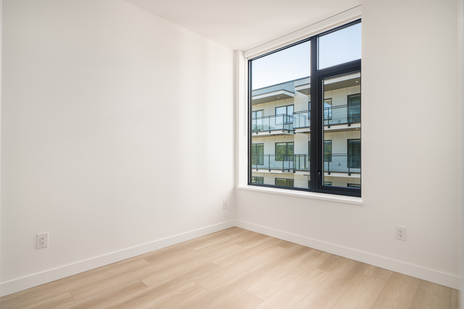 Unfurnished room with light wood flooring, white walls, and a large window showing a view of a modern apartment building with balconies.