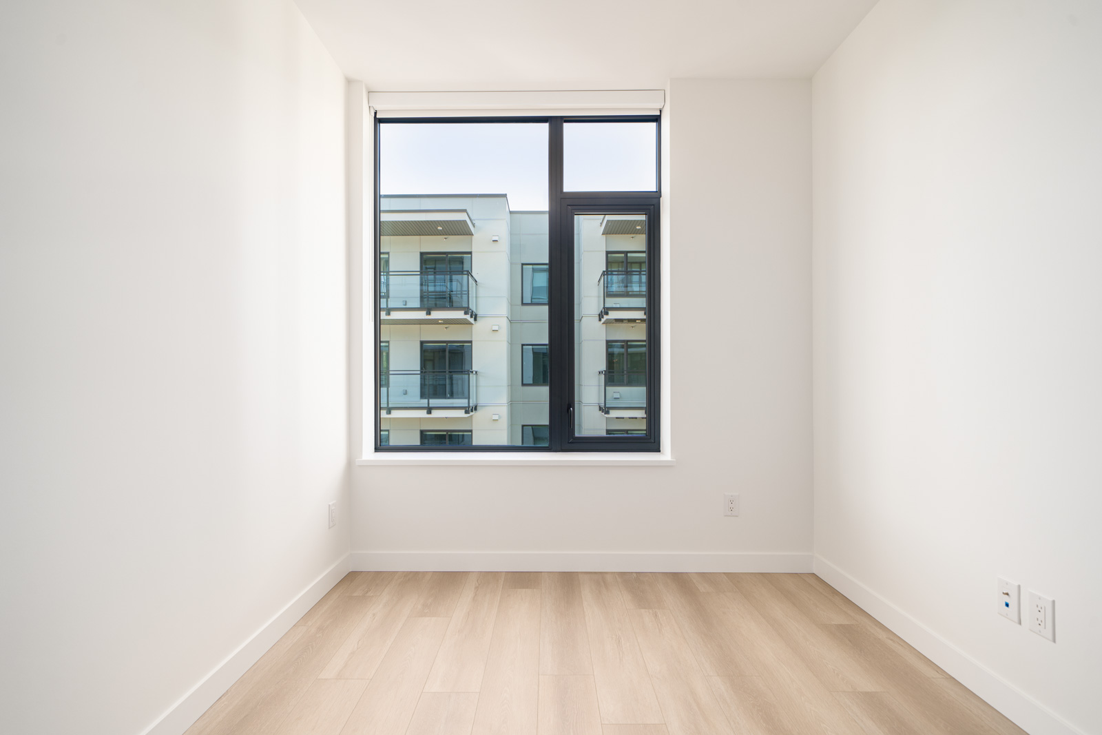 Empty room with light wood flooring, white walls, and a large window overlooking a modern apartment building with balconies.