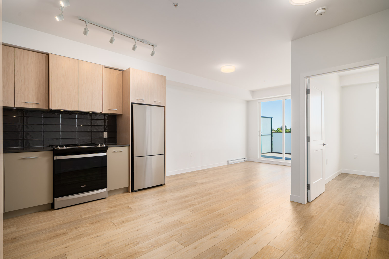 Modern apartment interior with light wood floors, a small kitchen area with stainless steel appliances, and an open living space leading to a balcony through glass doors.