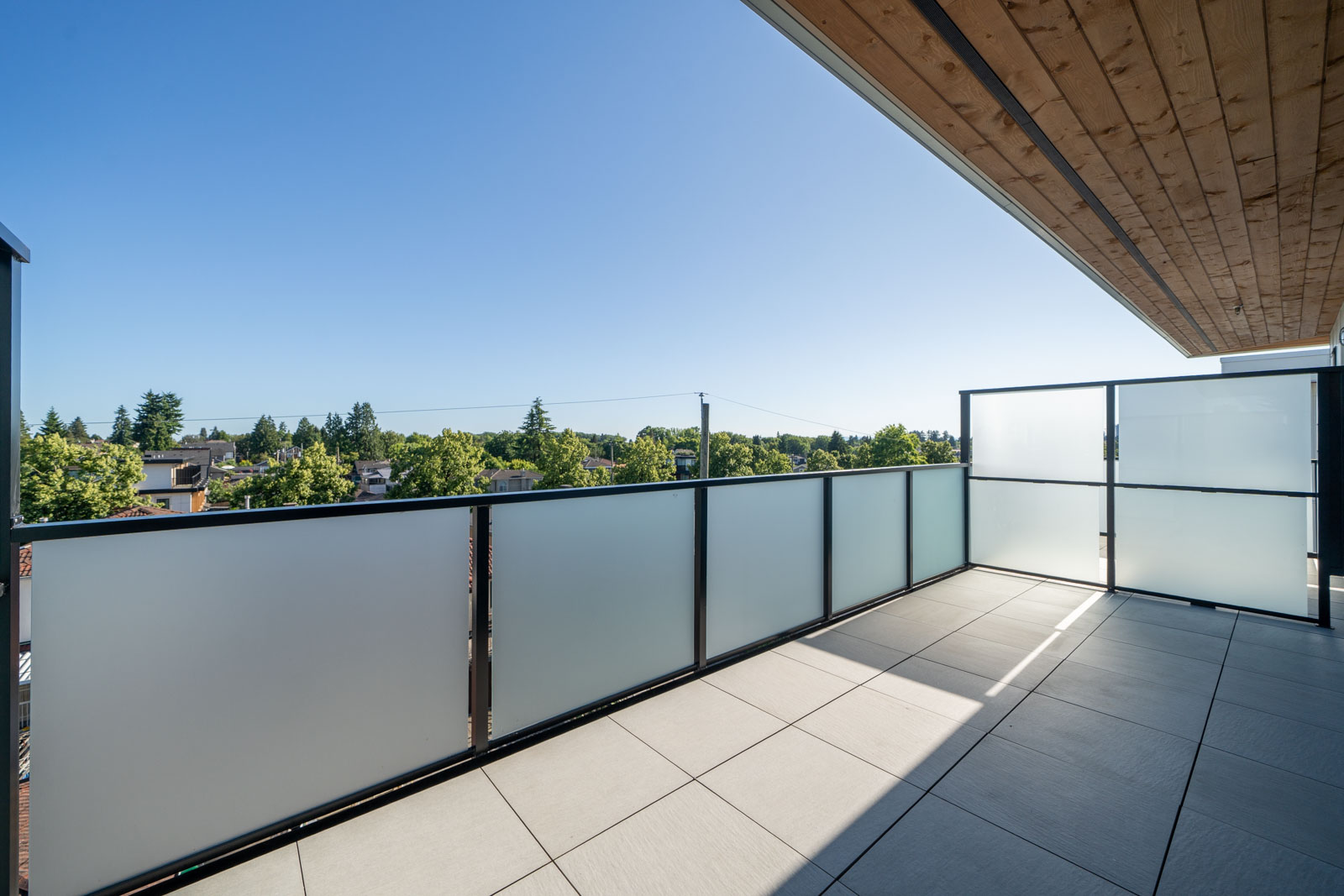 Spacious outdoor balcony with frosted glass railing, tiled floor, and partial wooden ceiling, overlooking trees and rooftops under a clear blue sky.