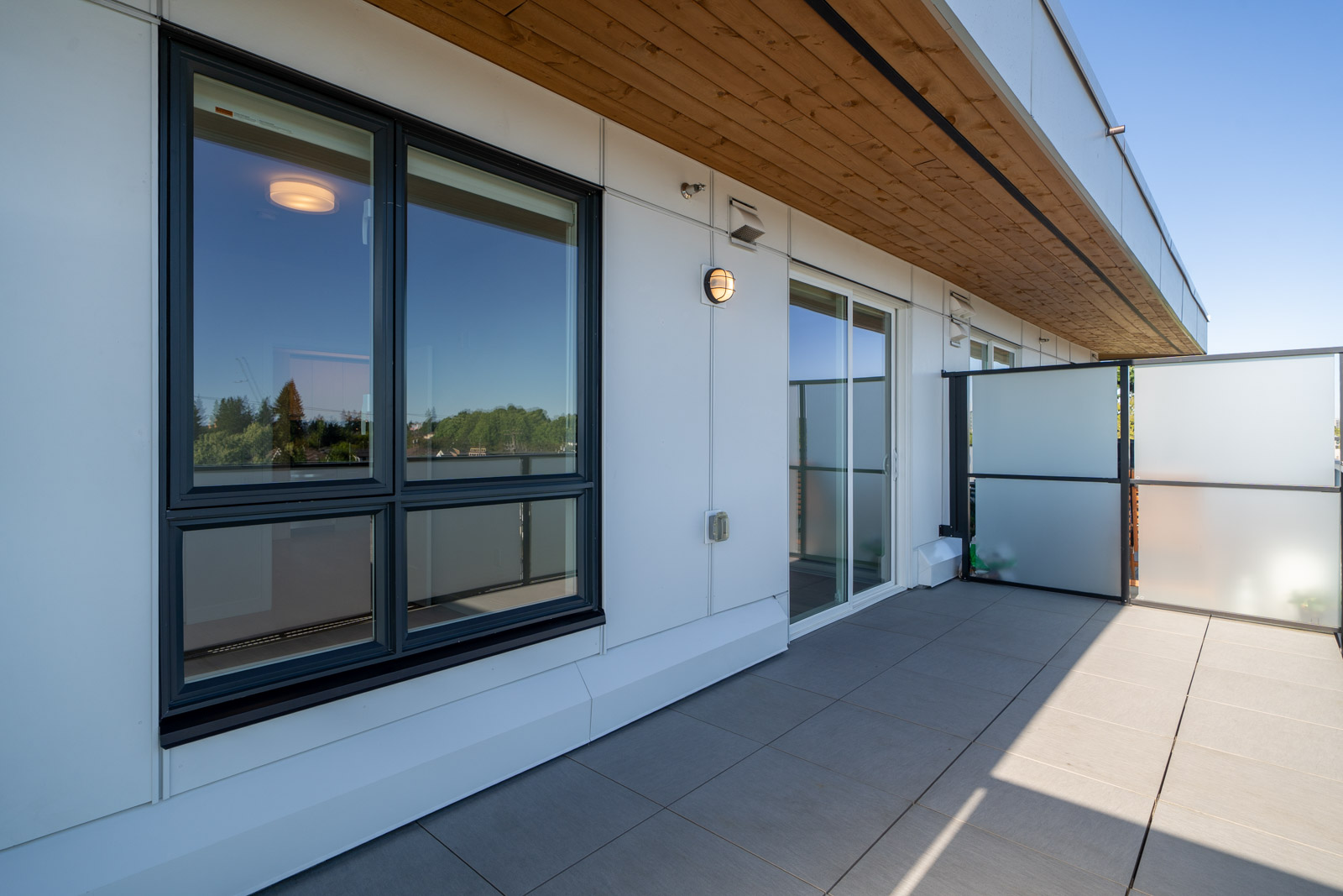 Modern apartment balcony with frosted glass privacy panels, large windows, tile flooring, and a wooden overhang under a clear blue sky.