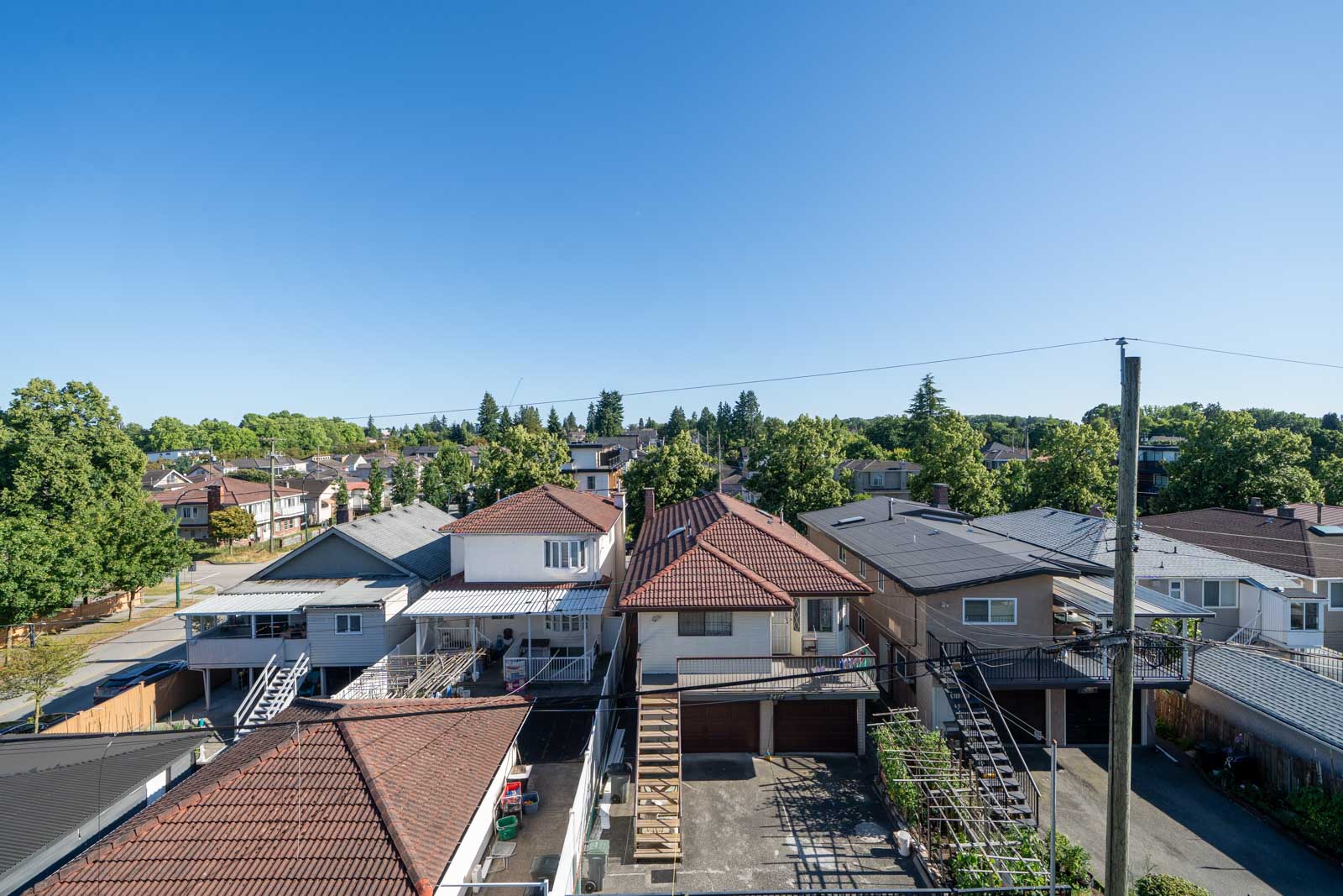 A row of suburban houses with red-tiled and grey roofs, driveways, and trees in the background under a clear blue sky.