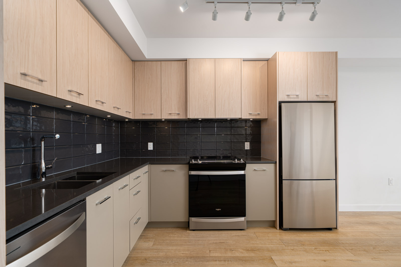 Modern kitchen with light wood cabinets, black tile backsplash, stainless steel appliances, and light wood flooring.