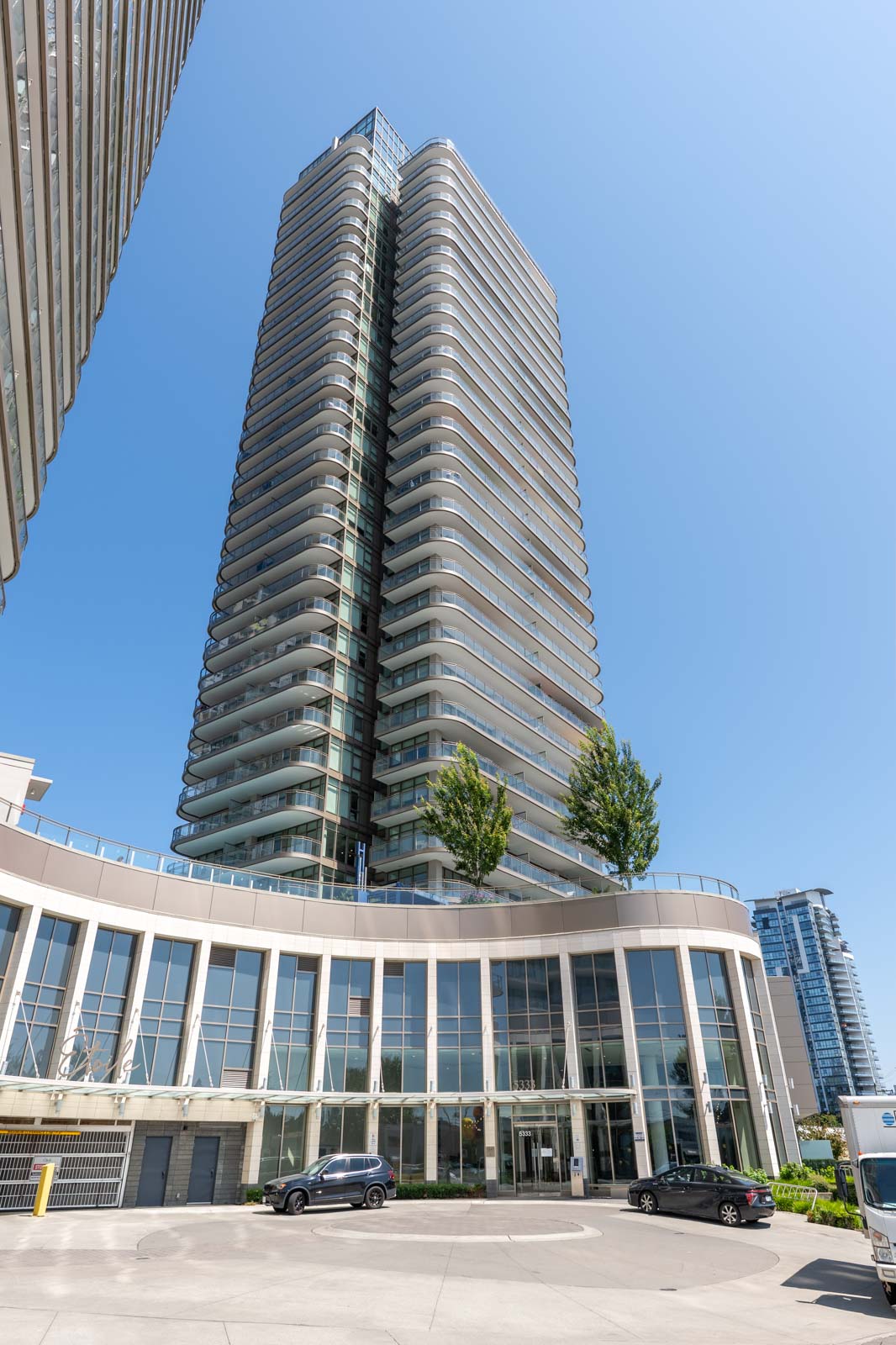 High-rise residential building with glass balconies and a curved lower facade, featuring trees on a terrace above the entrance, under a clear blue sky.