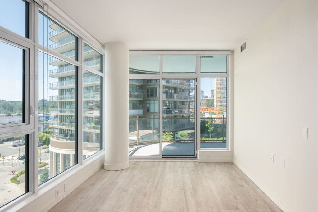 Unfurnished modern room with large floor-to-ceiling windows, light wood flooring, and a balcony overlooking nearby buildings.