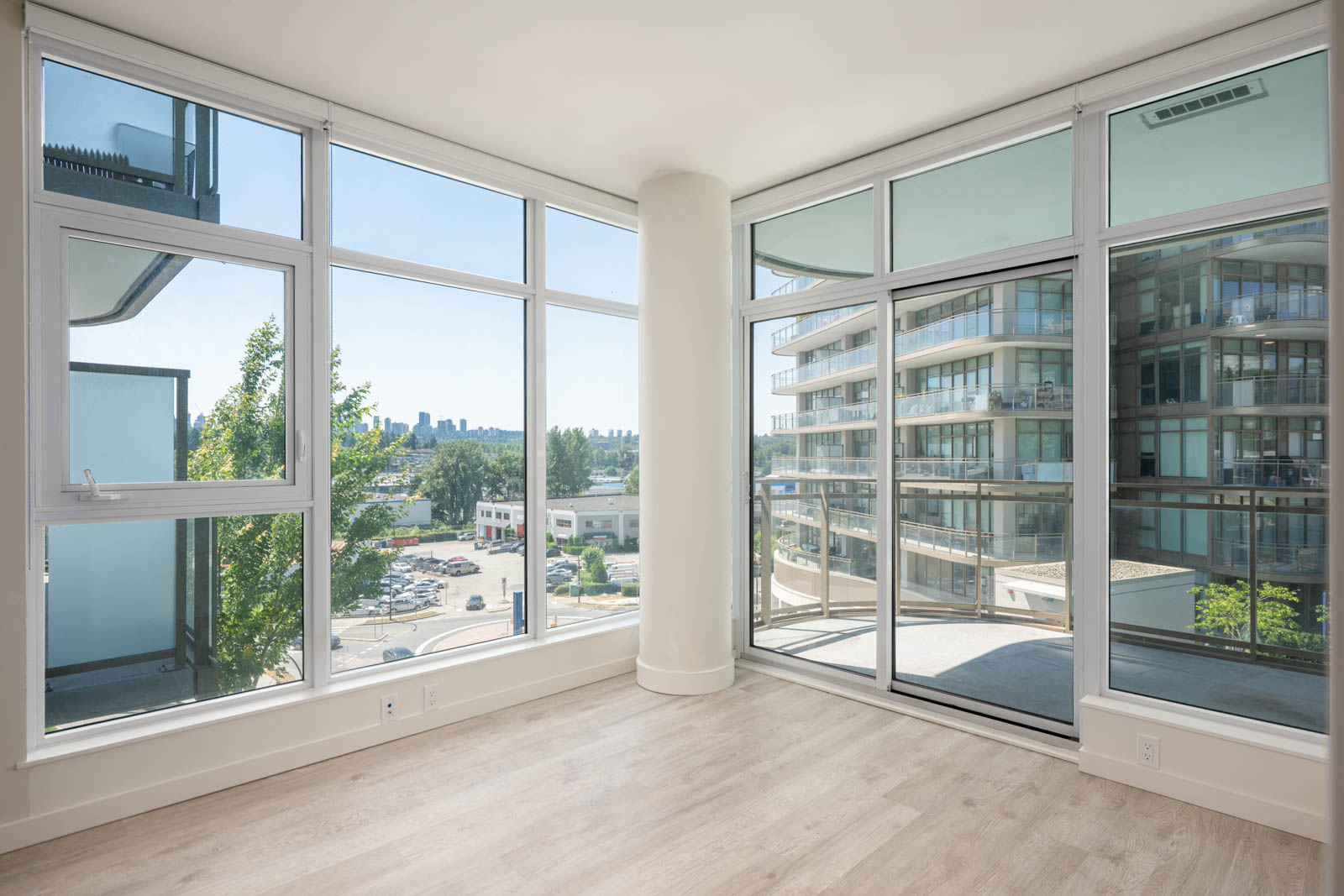 Unfurnished modern corner apartment with large floor-to-ceiling windows, light wood flooring, and a view of nearby buildings and cityscape.
