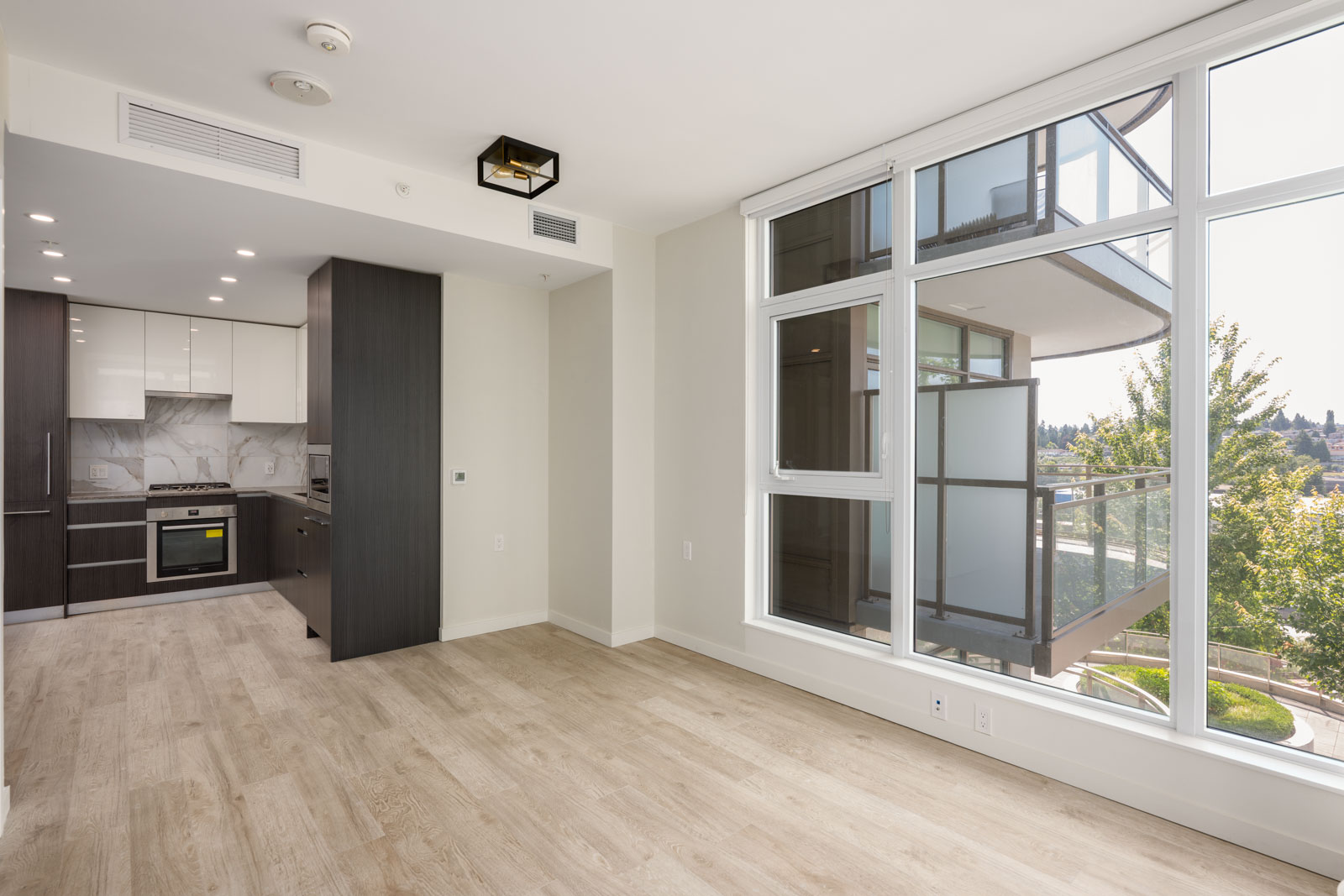 Modern apartment interior with light wood flooring, large windows, and an open kitchen featuring dark cabinets and stainless steel appliances; balcony and greenery visible outside.