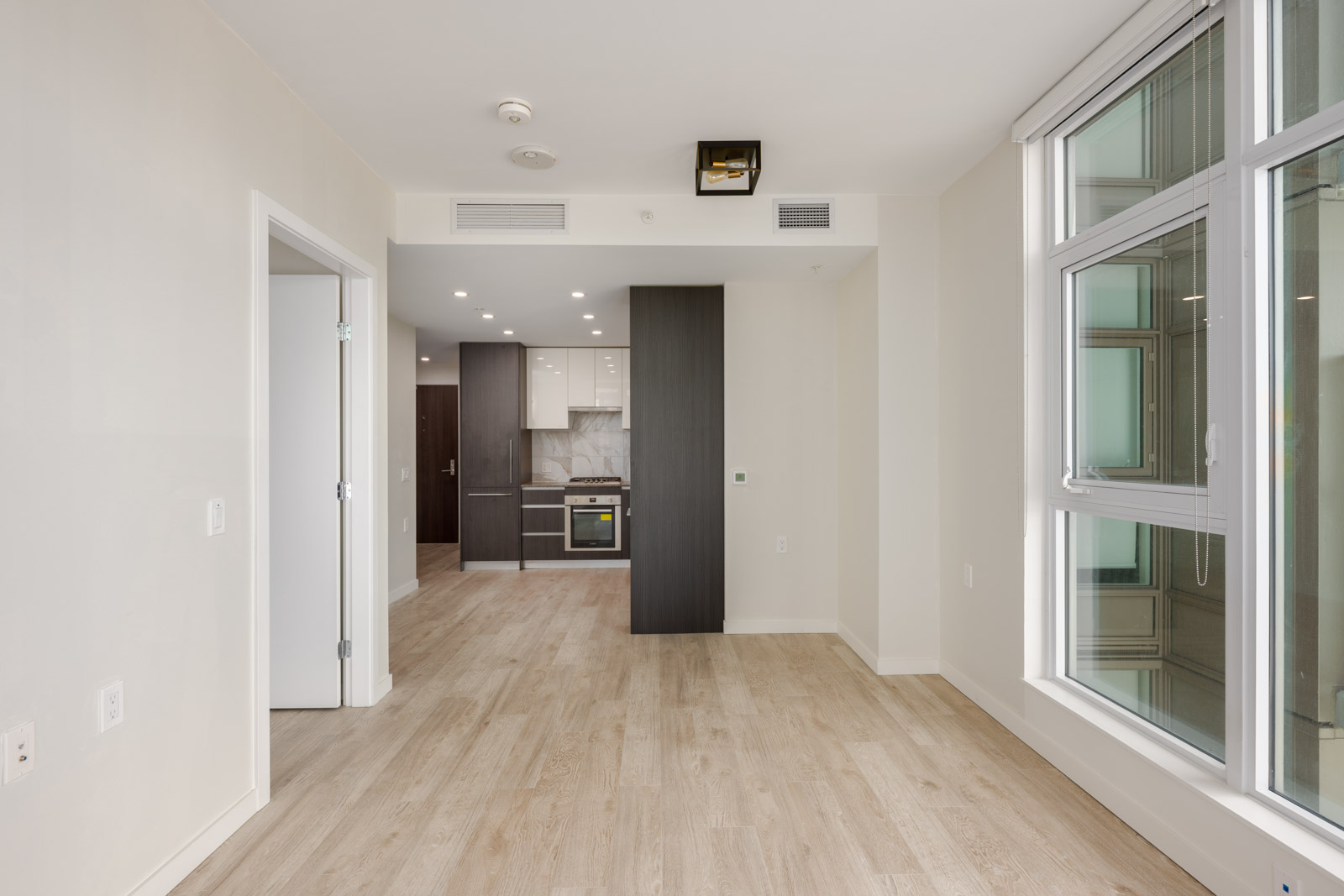A modern, unfurnished apartment with light wood flooring, white walls, large windows, and an open kitchen in the background.