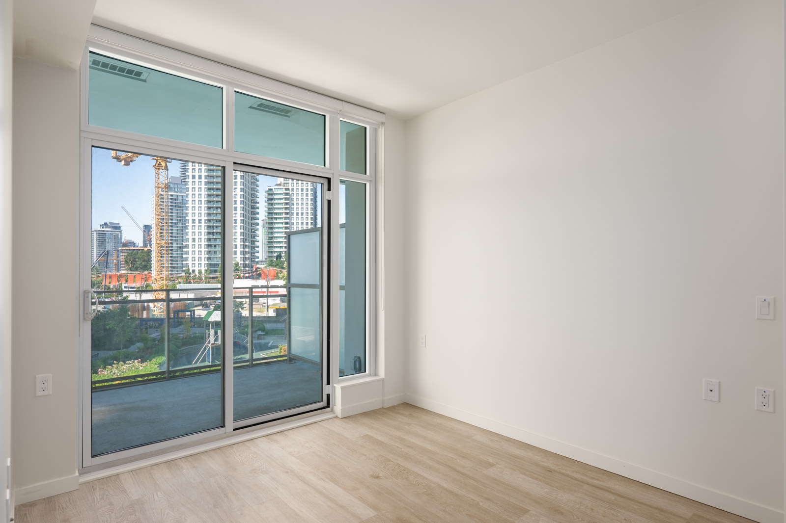 Unfurnished room with light wood flooring and large sliding glass doors leading to a balcony with a view of city buildings and construction cranes.