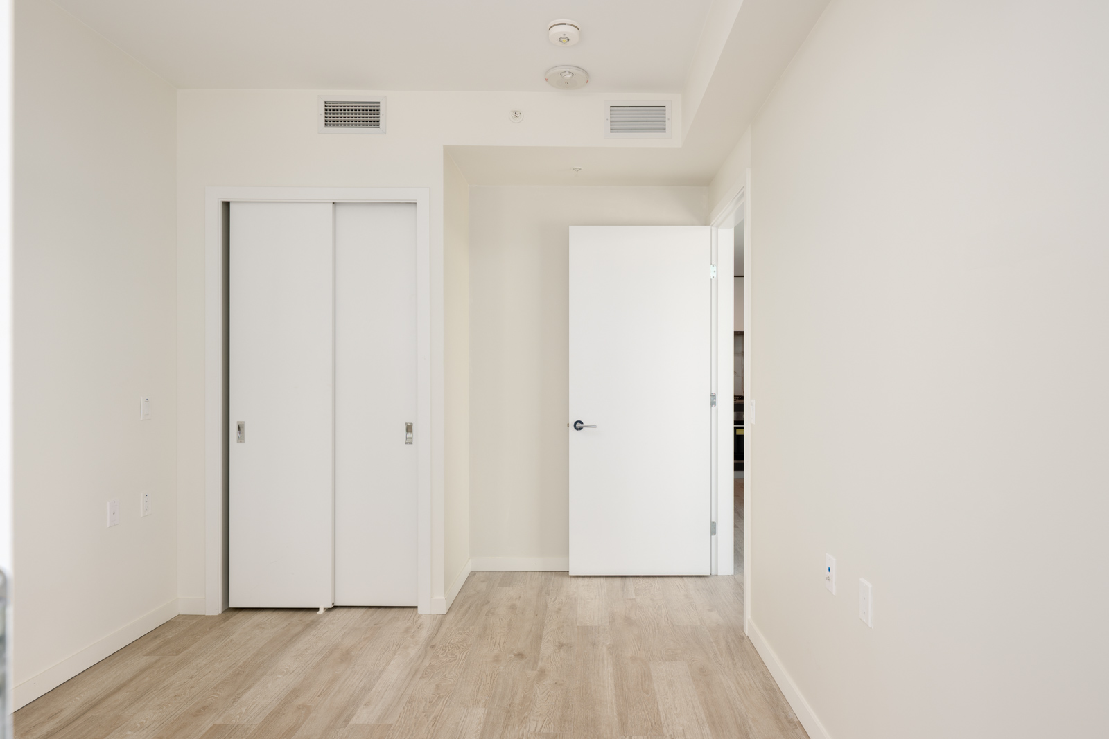 Empty room with light-colored walls, wood flooring, a white sliding-door closet, and an open white door leading to another area.