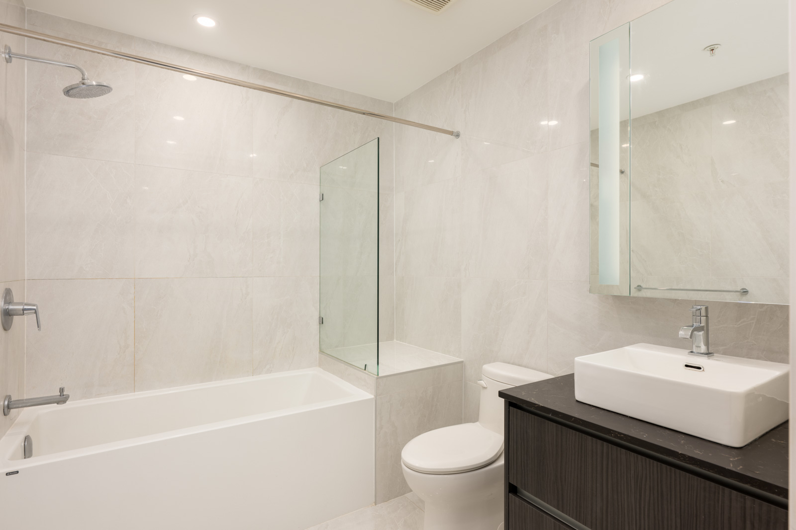 Modern bathroom with white tiles, a bathtub-shower combo with glass partition, toilet, rectangular sink on a dark vanity, and a wall mirror.
