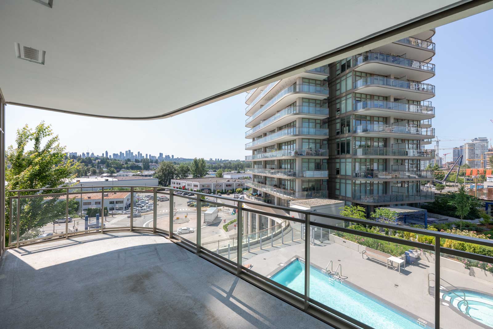 View from a curved balcony overlooking a swimming pool, nearby high-rise building, and a distant city skyline under a clear sky.