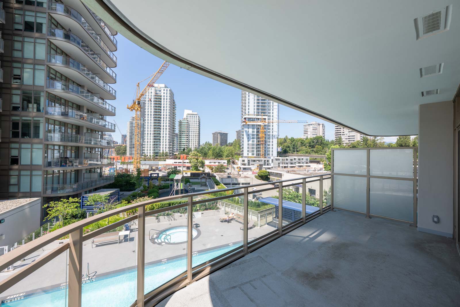 A view from a modern balcony overlooking a construction site, high-rise buildings, a swimming pool, and trees on a clear day.