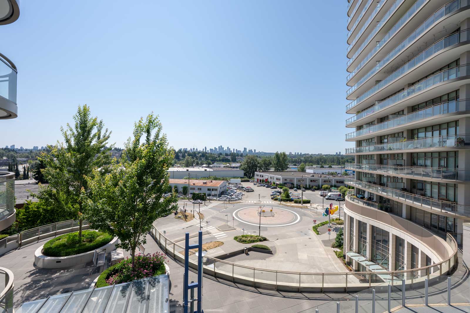 View of a modern urban plaza with trees, circular seating area, and high-rise buildings under a clear blue sky. City skyline visible in the background.