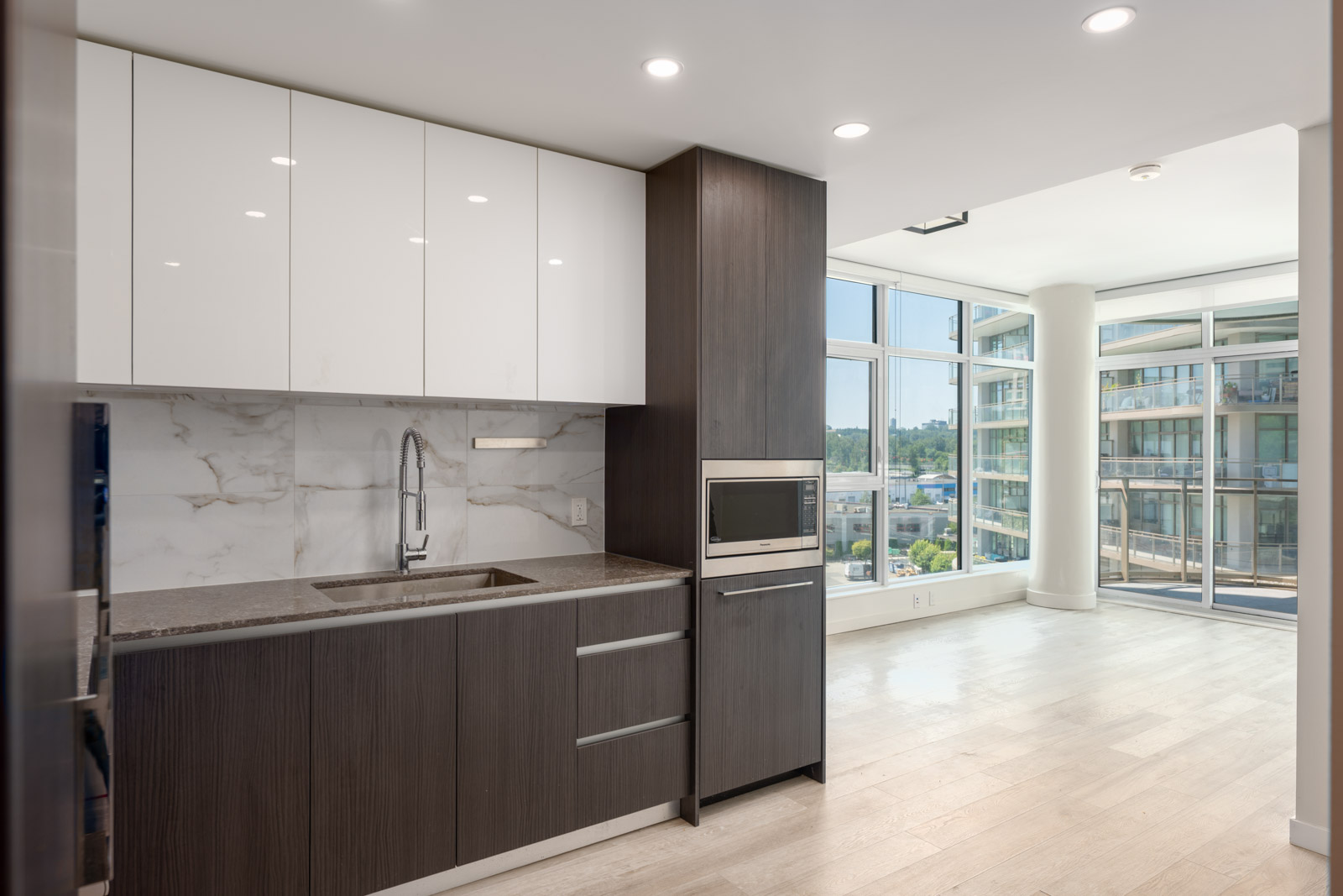 Modern kitchen with dark and white cabinets, marble backsplash, built-in microwave, and large windows overlooking a bright living area and cityscape.