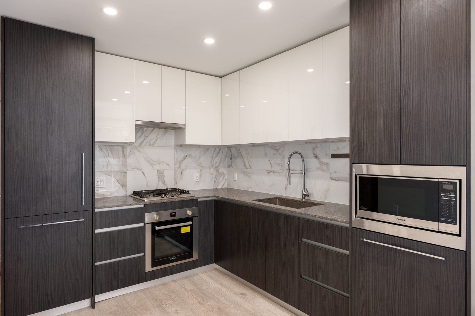 Modern kitchen with dark wood and white cabinets, built-in oven, stovetop, microwave, marble backsplash, and a stainless steel sink under bright recessed lighting.