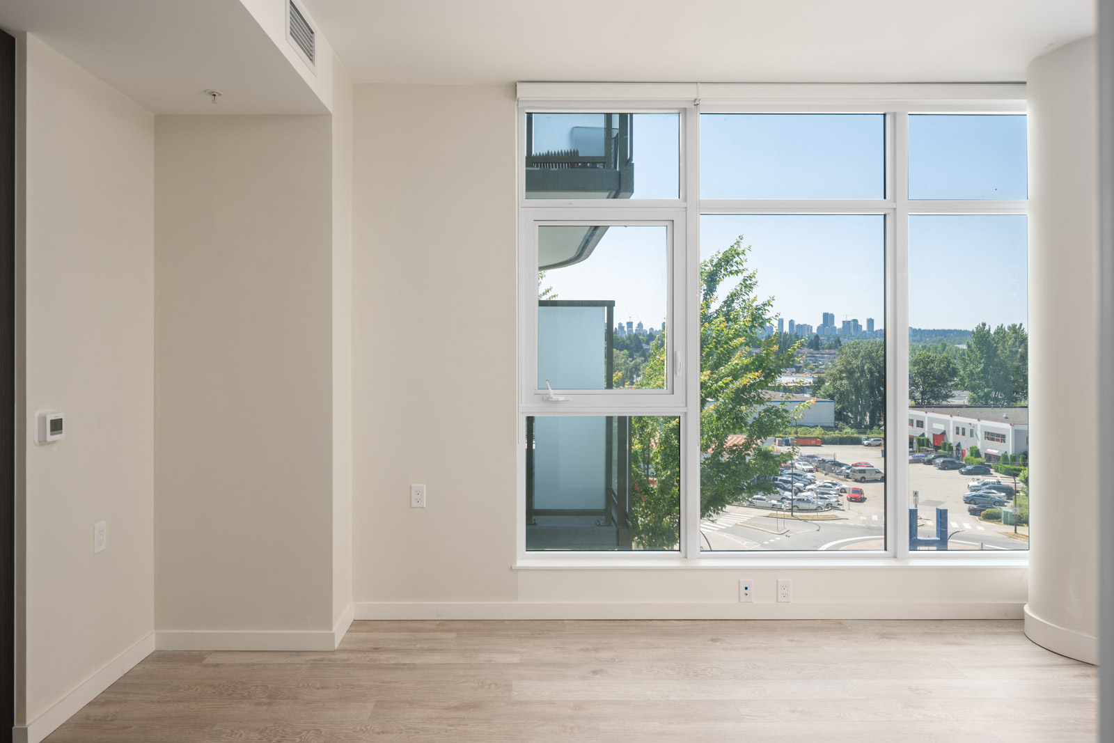 Empty room with light-colored walls and wood flooring, featuring a large window overlooking a parking lot, trees, and distant city buildings.