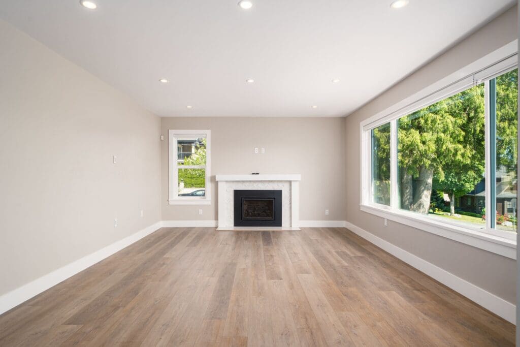 Empty living room with light-colored walls, wood flooring, a fireplace, recessed ceiling lights, and large windows showing trees and a house outside.