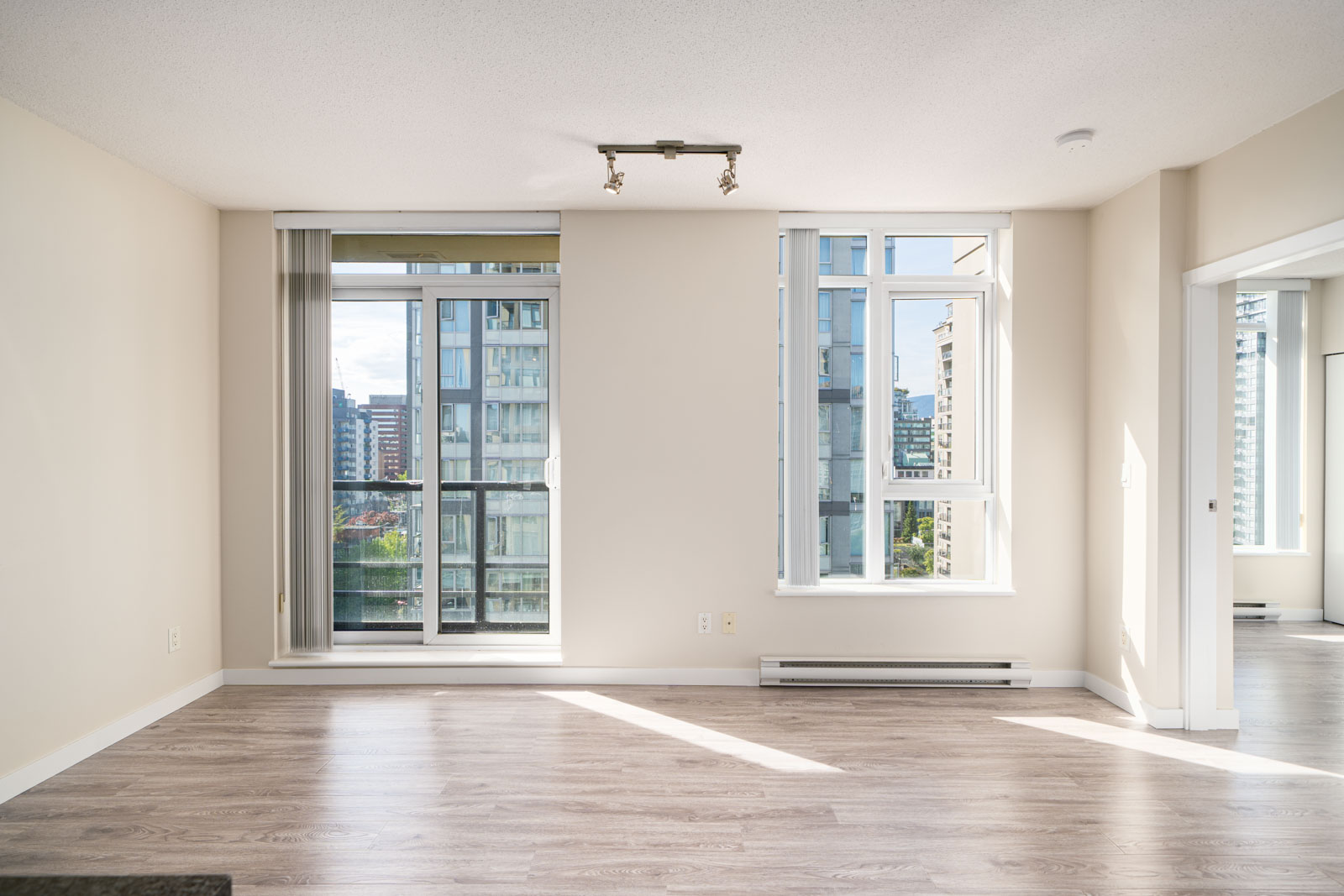 Unfurnished apartment room with light wood flooring, large windows, and a glass door opening to a balcony, with city buildings visible outside.