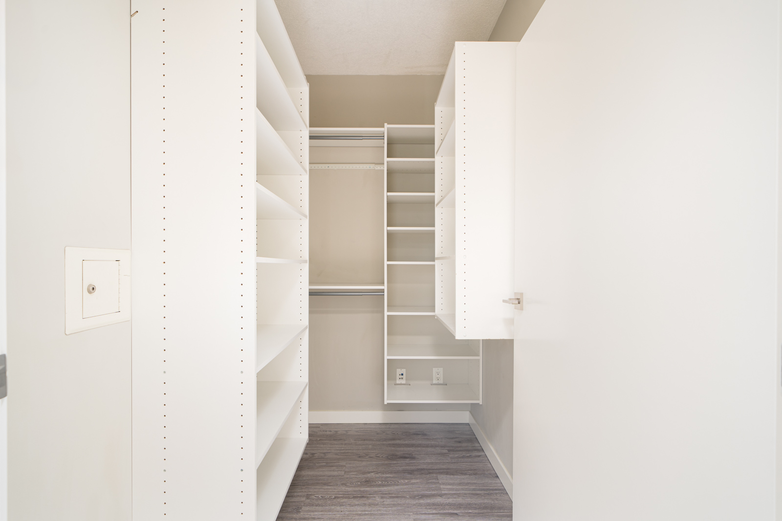 Empty walk-in closet with white shelving units, open cabinets, and hanging rods, featuring light-colored walls and dark wood flooring.