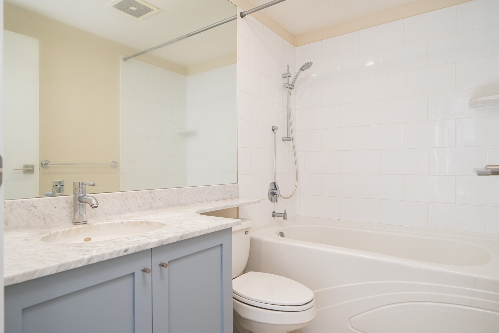 Bathroom with a marble countertop, blue vanity, toilet, large mirror, and a bathtub with a showerhead against a tiled wall.