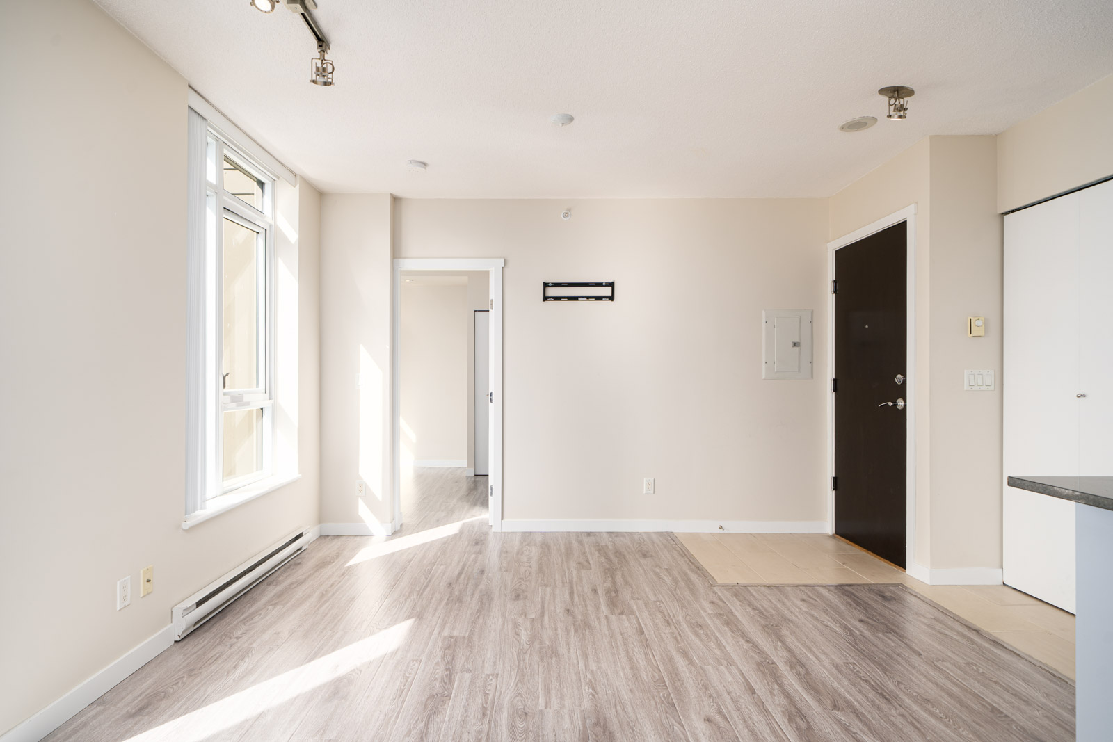 Bright, empty apartment interior with light wood flooring, large window, beige walls, and a black entrance door. Doorway leads to another room.