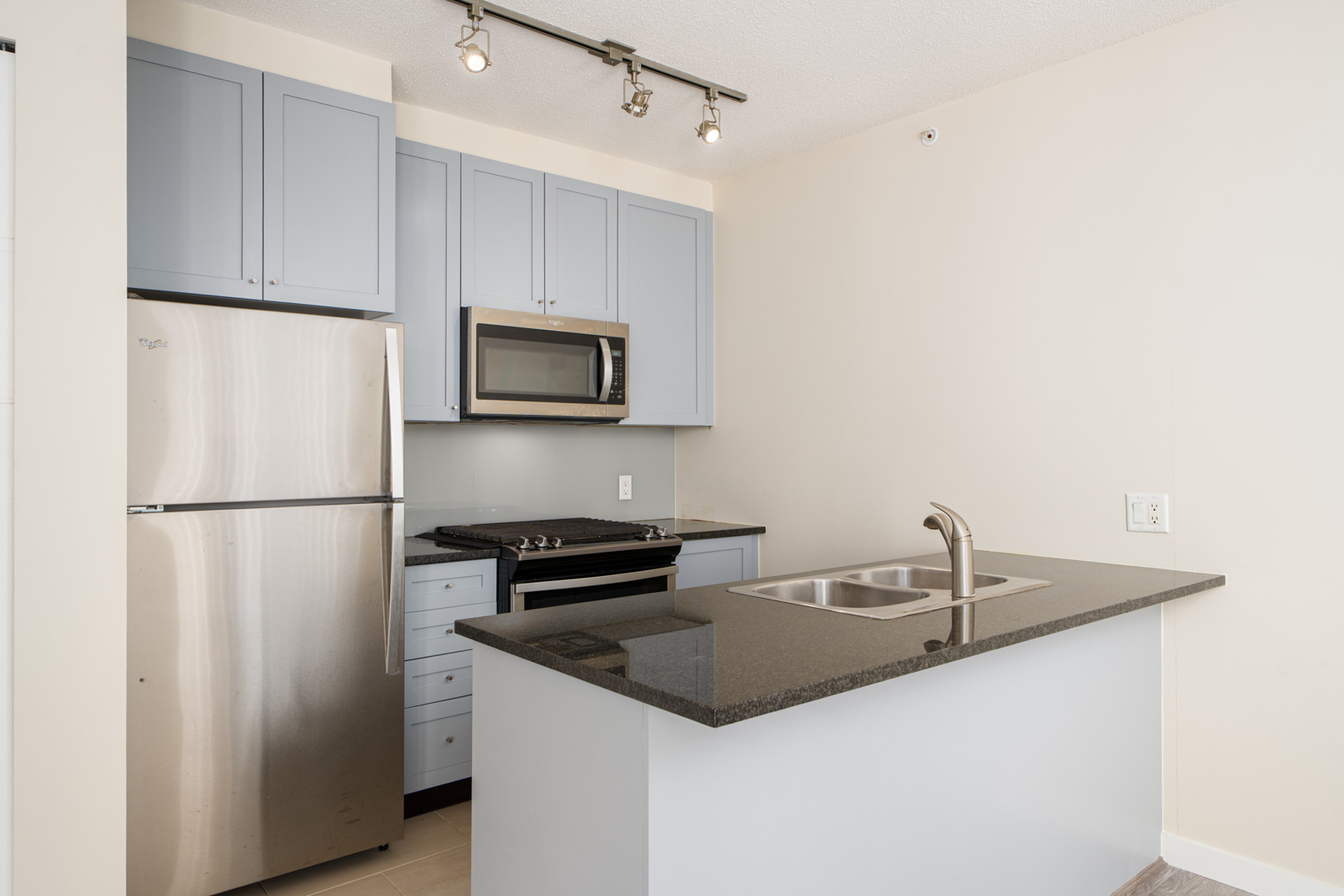 Modern kitchen with light blue cabinets, stainless steel refrigerator, built-in microwave, oven, and a black granite island with a double sink and chrome faucet.