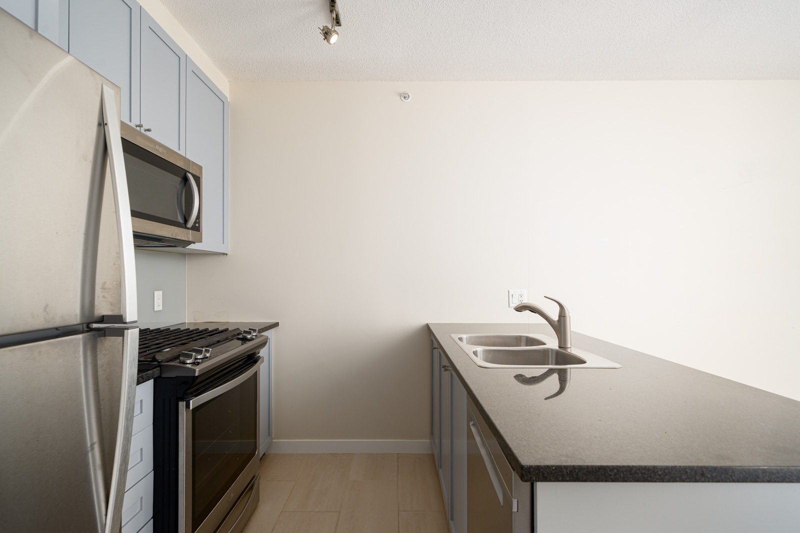 Modern kitchen with stainless steel appliances, double sink, black countertop, light blue cabinets, beige floor tiles, and plain white walls.