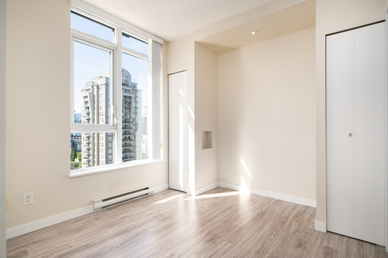 Bright empty room with large window, white walls, light wood flooring, two closets, and a cityscape view with high-rise buildings outside.