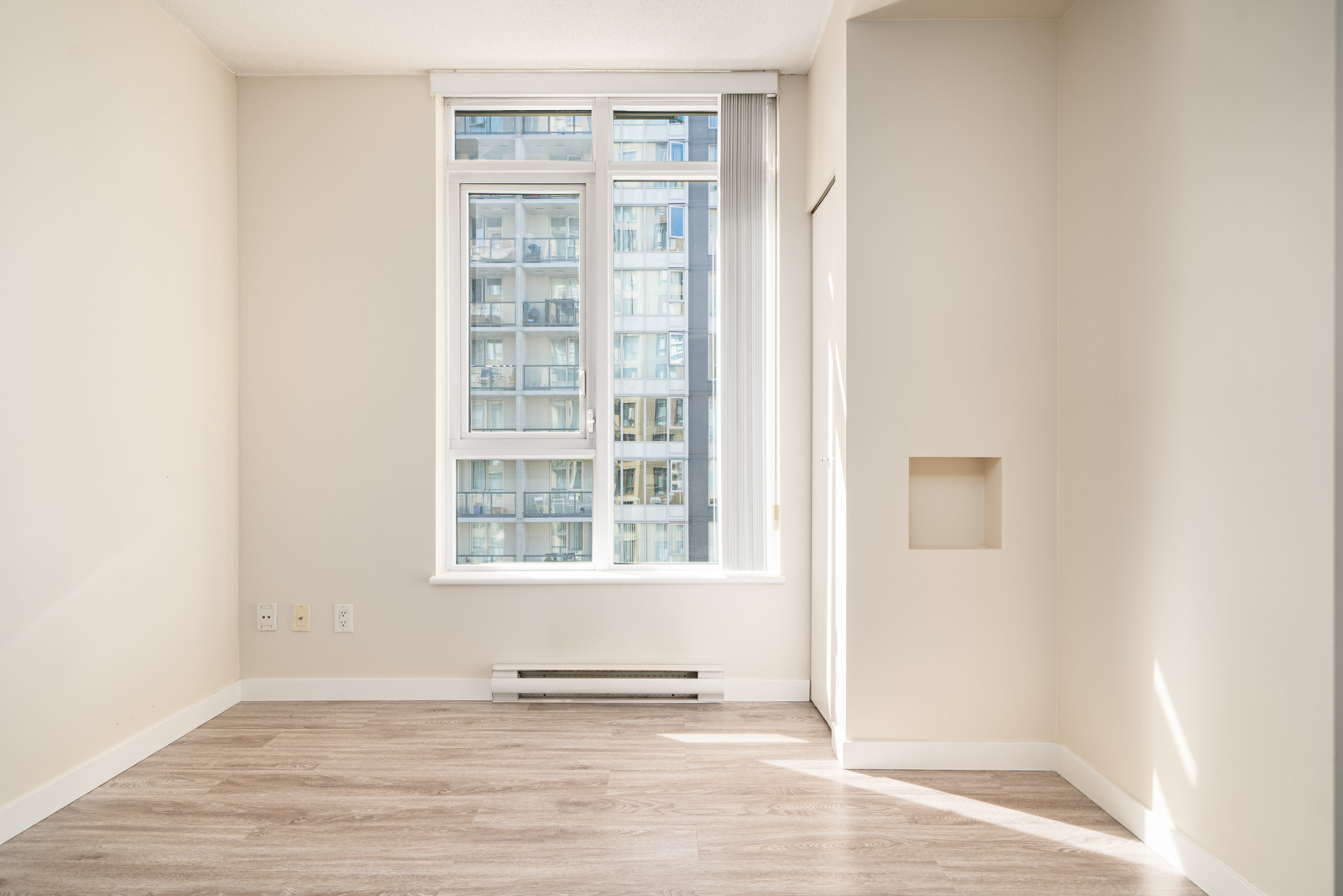 Bright, empty room with light wood flooring, cream-colored walls, and a large window showing a neighboring modern glass building.