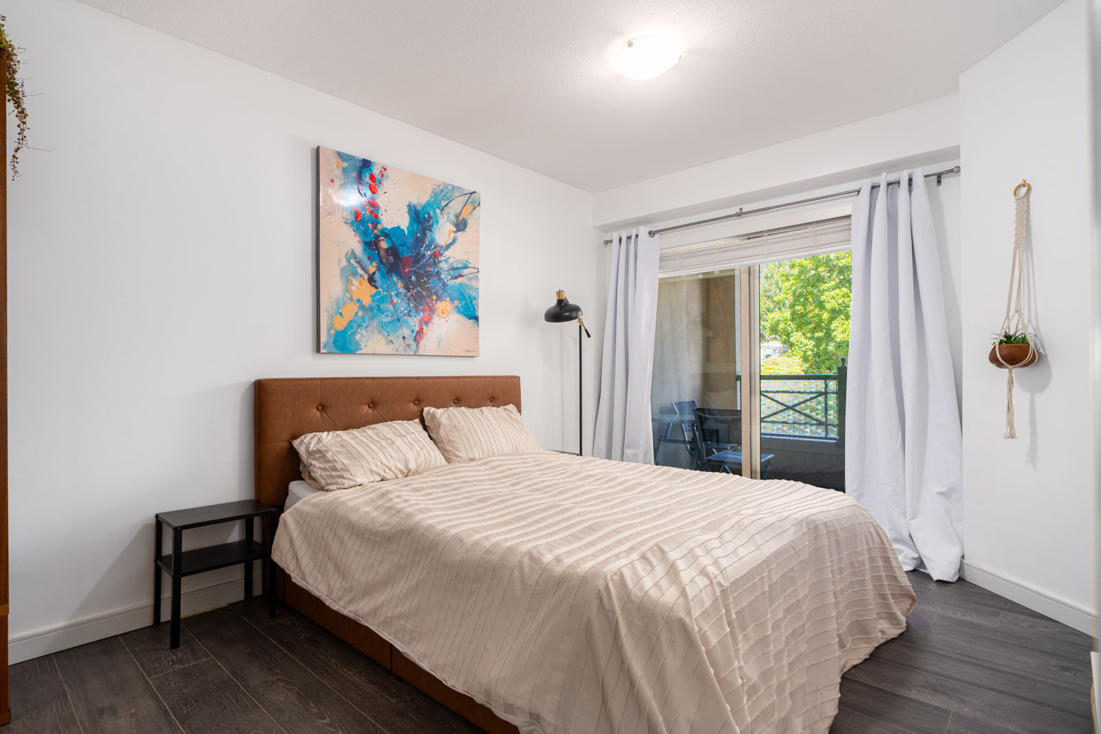 A neatly made bed with beige bedding sits against a white wall, under an abstract painting. A window with curtains and a sliding door opens to a balcony with greenery outside.