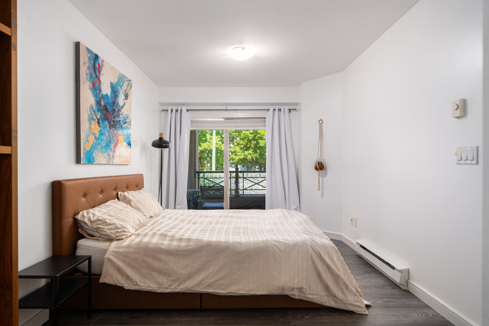 A modern bedroom with a neatly made bed, abstract artwork above the headboard, white walls, and a window with curtains overlooking a balcony with greenery outside.