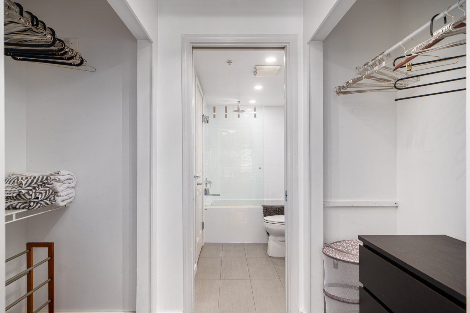 View of a modern bathroom with a bathtub and toilet, seen from a walk-in closet with empty shelves, hangers, and folded towels.