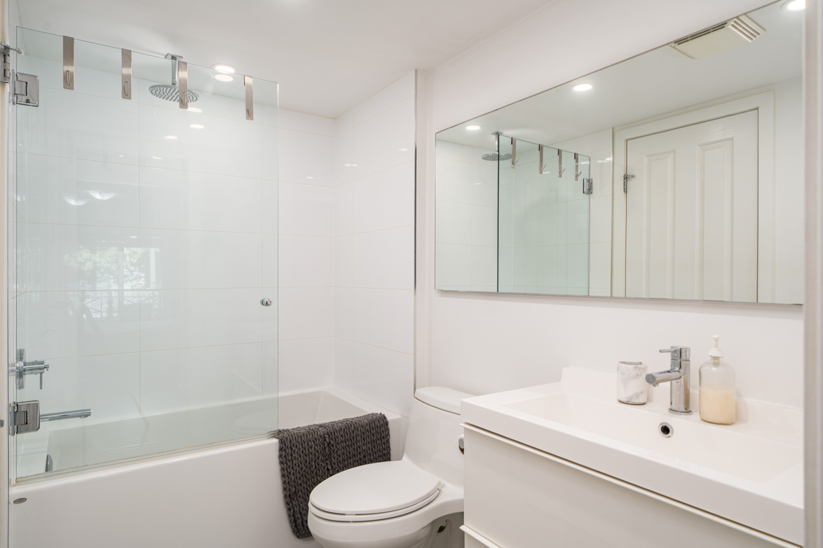 Modern white bathroom with a bathtub-shower combo, a toilet, a white vanity with sink, and a large mirror above the sink. A dark towel hangs over the bathtub edge.