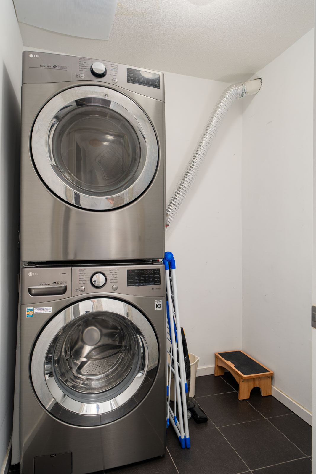 A stacked stainless steel washer and dryer in a laundry room with a vent hose, folding drying racks, and a small wooden step stool nearby.