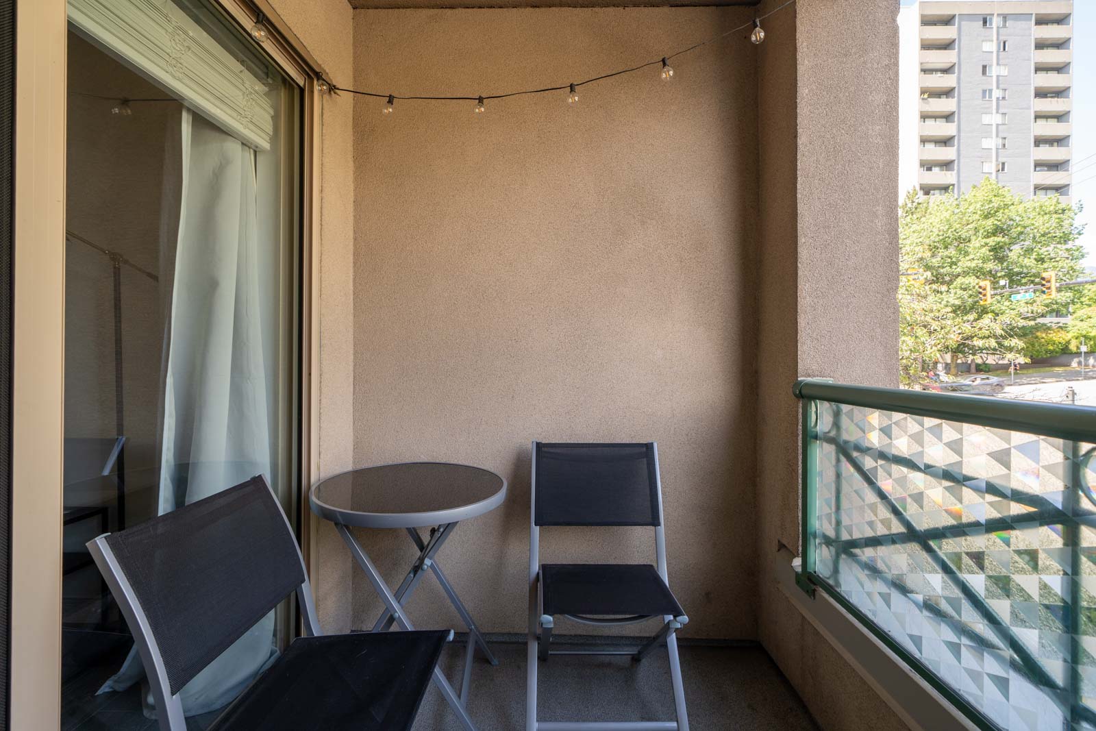Small balcony with two black chairs, a round table, string lights above, and a partial view of a neighboring building and trees in the background.