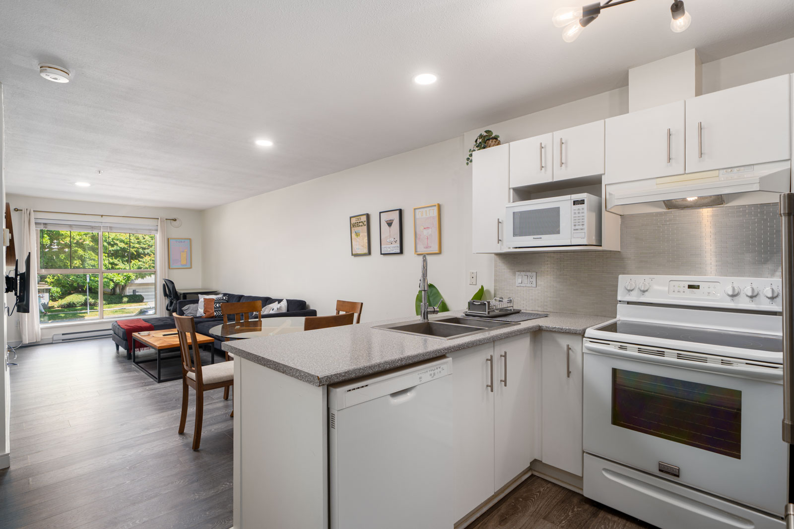 Modern open-concept kitchen with white cabinets and appliances, adjoining dining and living area with large window overlooking greenery.