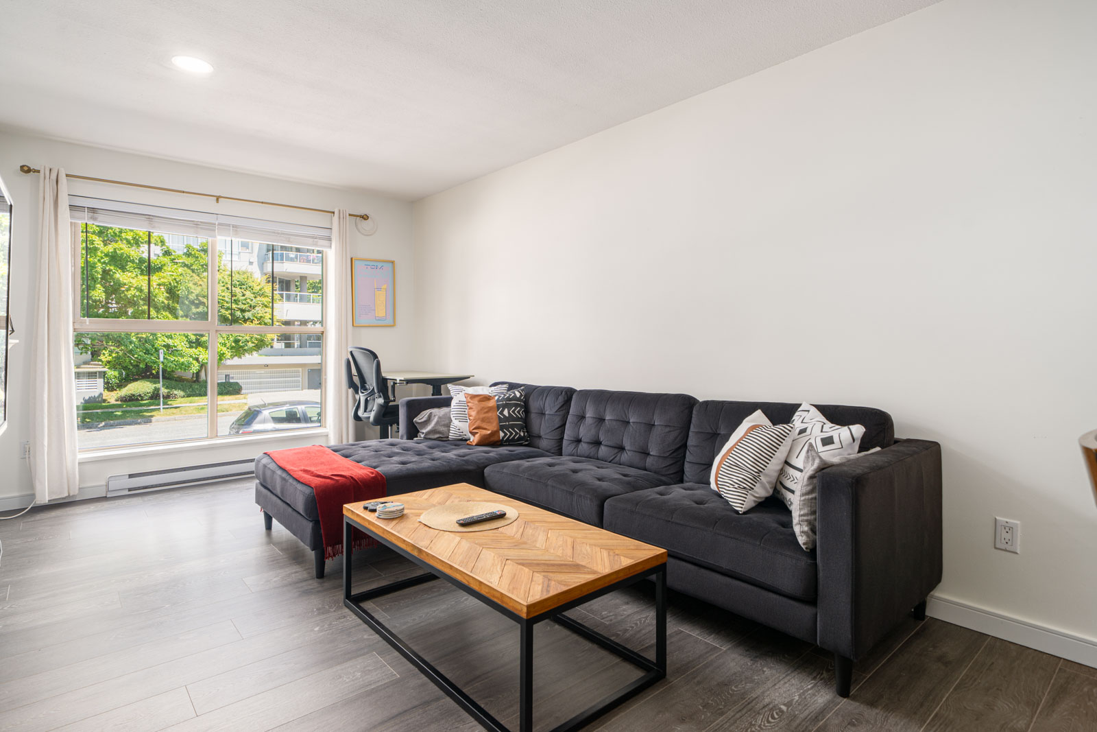 Modern living room with a large black sectional sofa, patterned pillows, wood coffee table, and window with daylight streaming in, overlooking trees and a street outside.