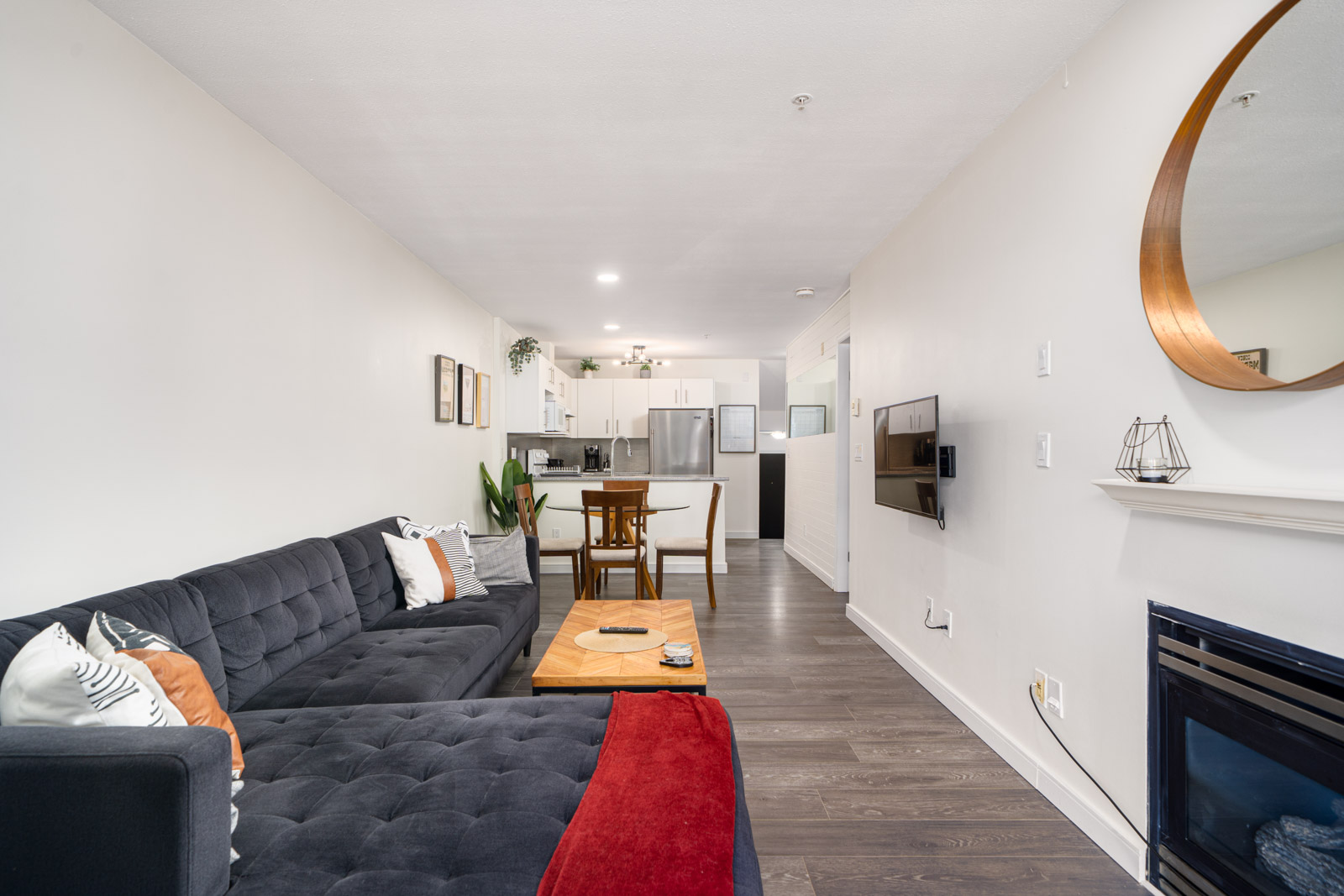 Modern living room with dark sectional sofa, wooden coffee table, wall-mounted TV, round mirror above fireplace, and open kitchen with dining area in the background.