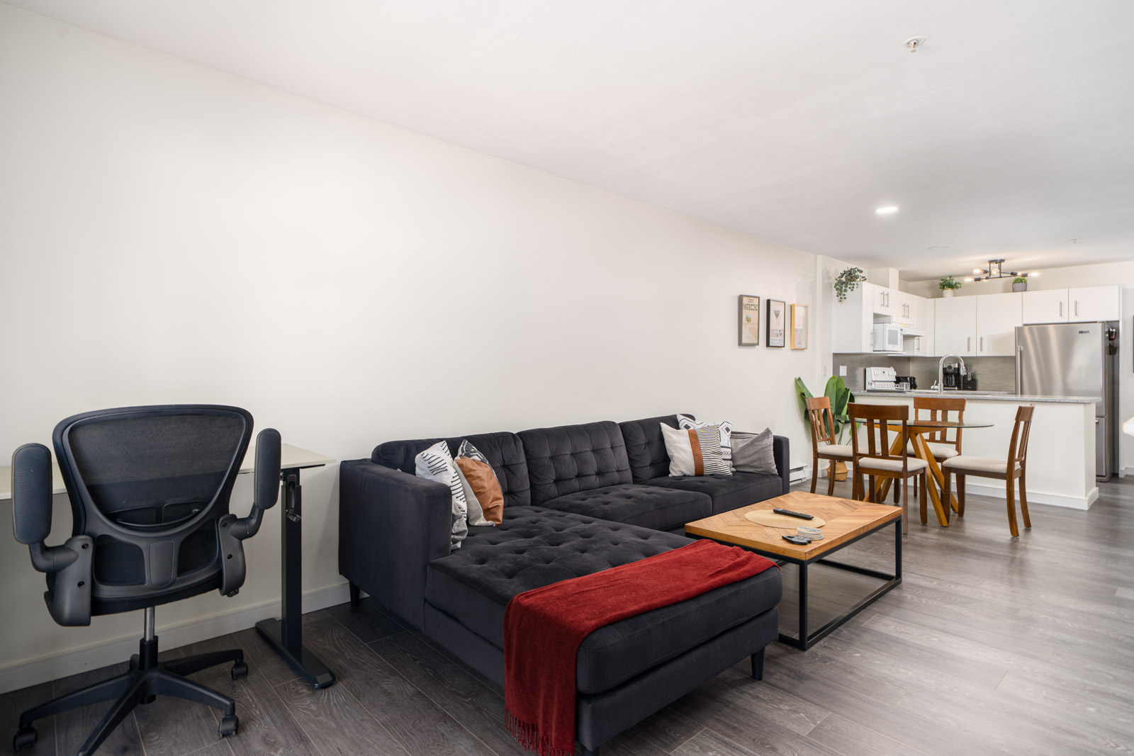 Modern living room with a black sectional sofa, desk and chair, wooden coffee table, dining table, and open kitchen in a bright, minimalist apartment.