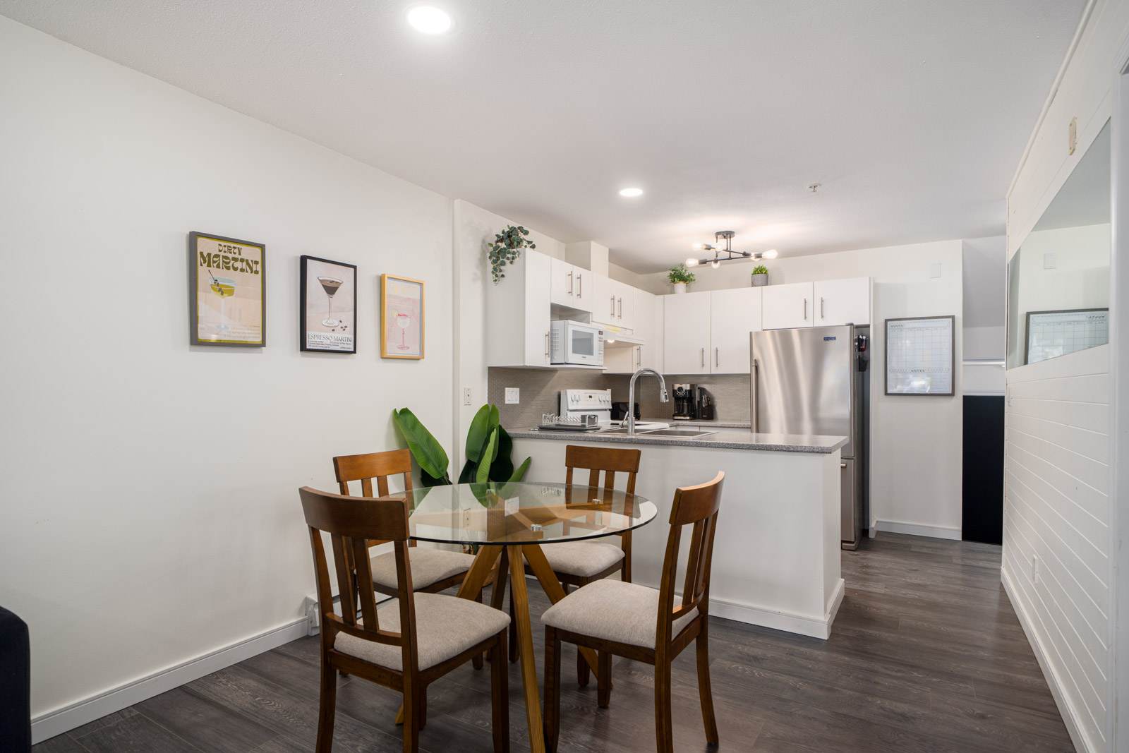 Modern kitchen and dining area with white cabinets, stainless steel appliances, a glass-top table with four wooden chairs, and framed art on the wall.