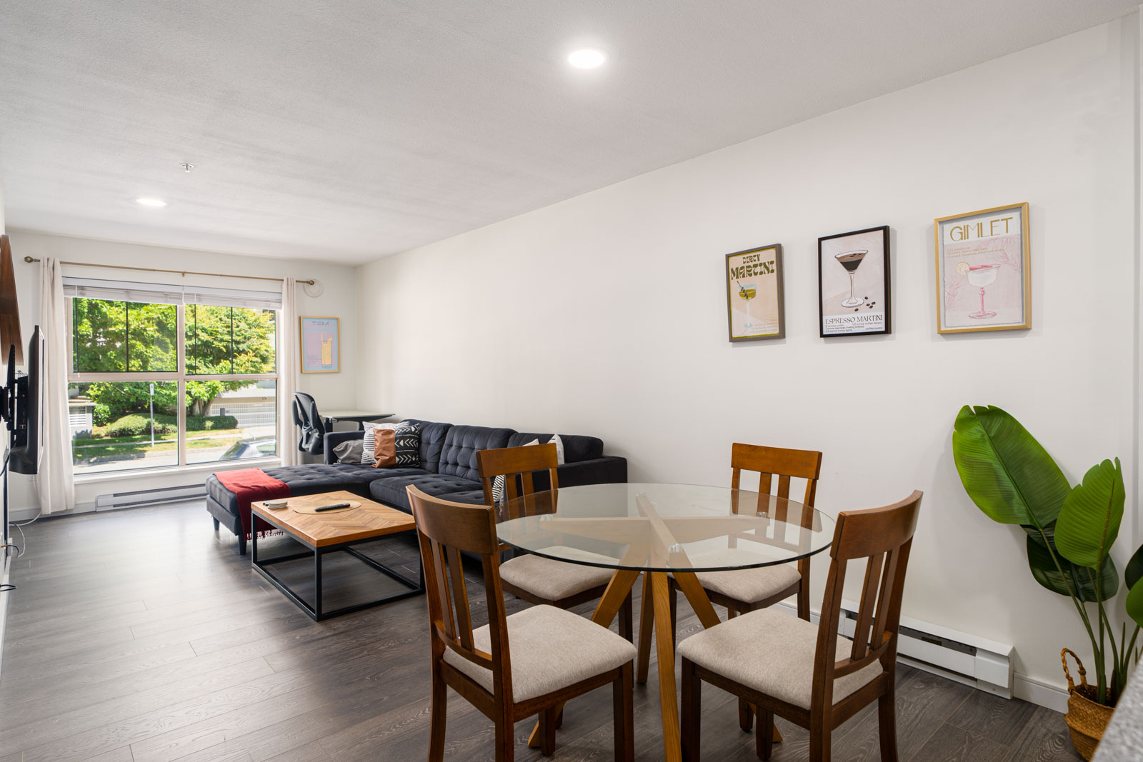 Modern living and dining area with a black sectional sofa, glass dining table with four chairs, wall art, large window, and a green potted plant.
