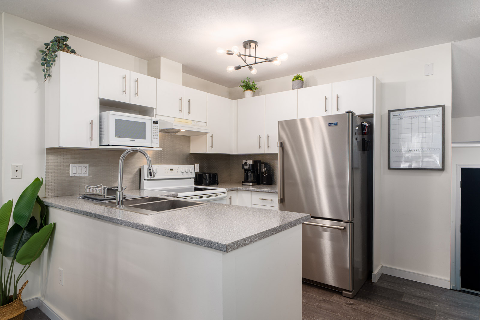 Modern kitchen with white cabinets, stainless steel appliances, a microwave, and potted plants on top of cabinets. A wall calendar and large plant are visible near the counter.