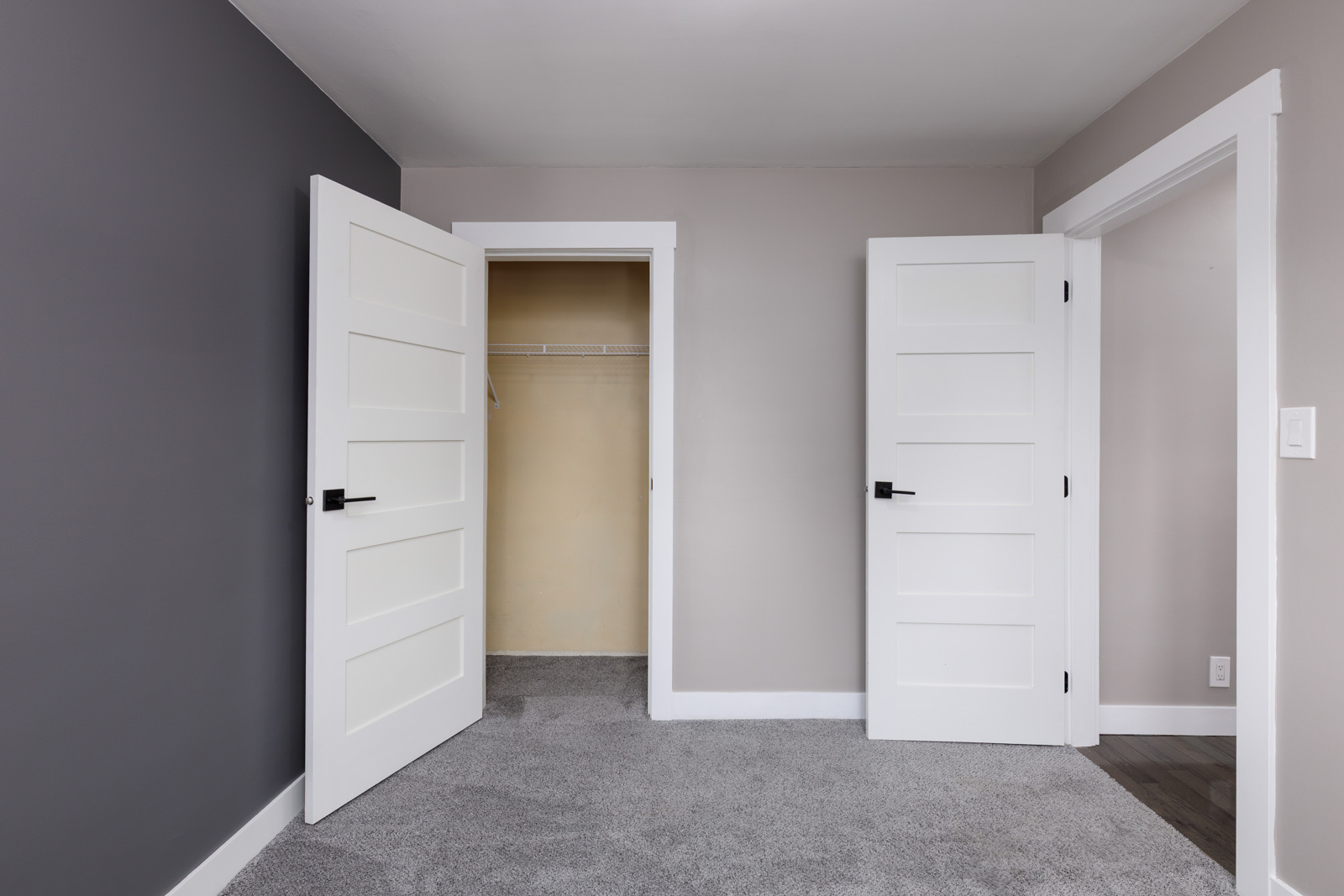 Empty room with gray carpet, one gray accent wall, two white doors; one door opens to a closet with a shelf and hanging rod, the other opens to a hallway.