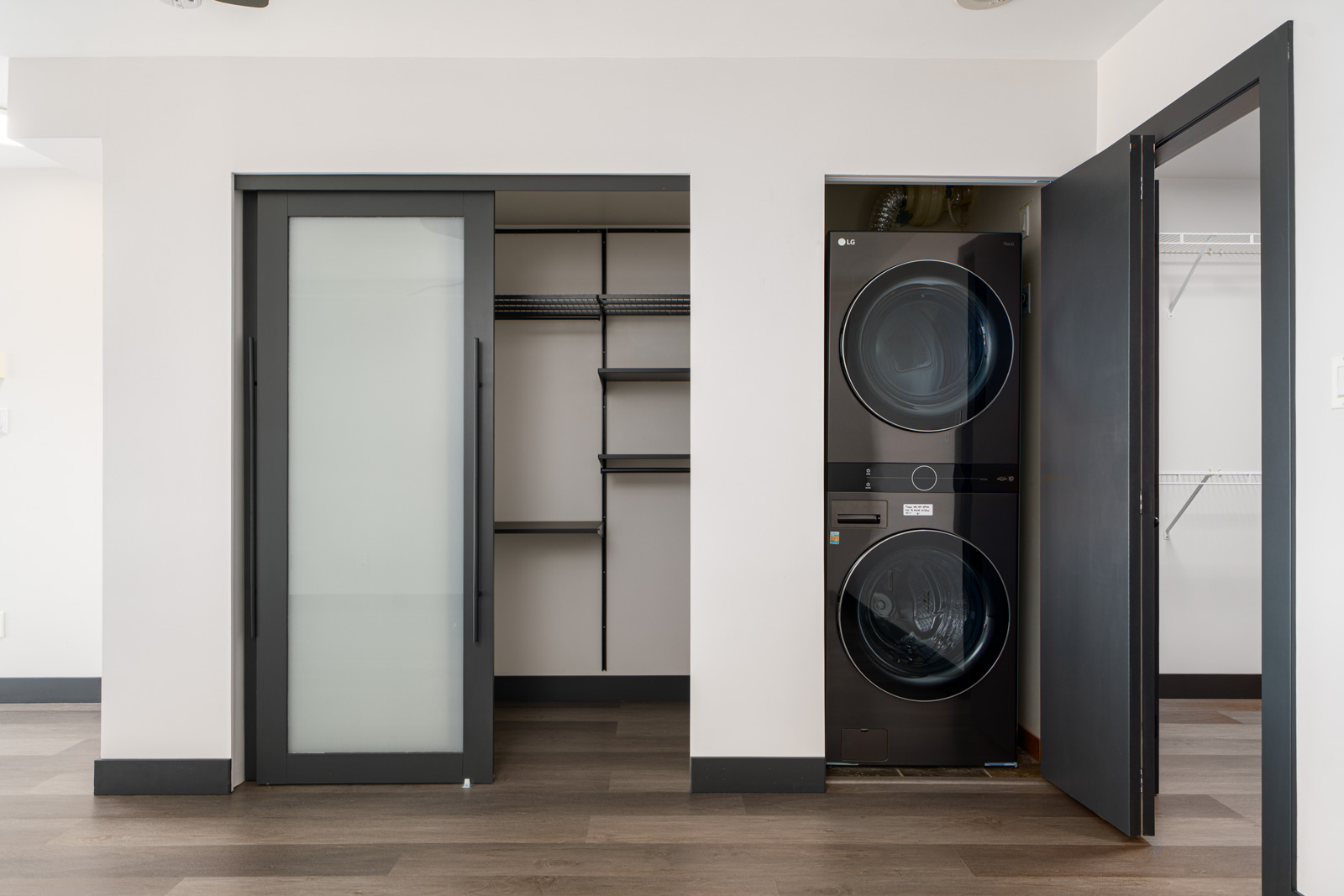 A modern laundry area with black stacked washer and dryer, open black closet doors, shelving, and wood-look flooring.